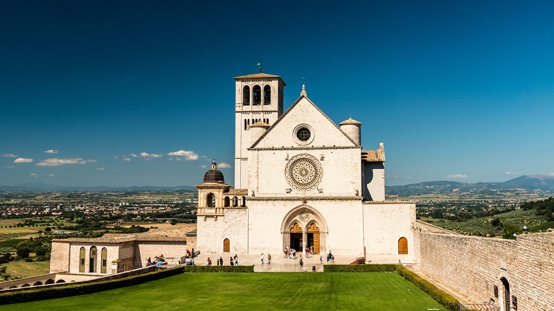 Franciscusbasilikan i Assisi lyser vitt i starkt solljus, kontrasterande mot en gr�n gr�smatta och bl� himmel.