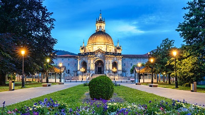 Santuario de Loyola i skymningen, med tända lyktor och upplyst kupol samt en blomsterrabatt i förgrunden.