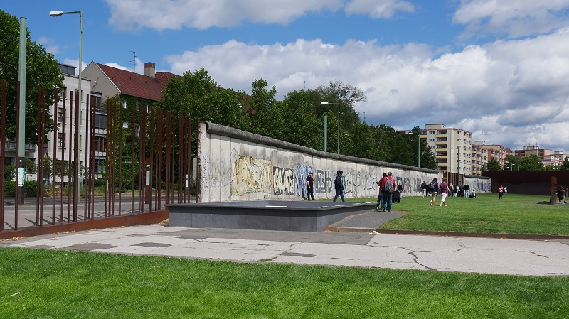 Del av Berlinmuren som m�lats med graffiti och m�nniskor som str�var omkring p� gr�set i n�rheten.